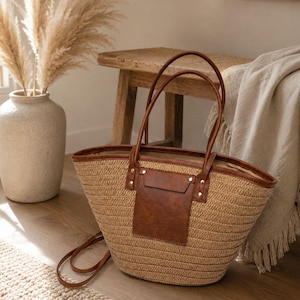 May include: A woven straw tote bag featuring brown faux leather trim, two long shoulder straps, and a rectangular brown faux leather front pocket secured with metallic rivets. The bag has a wide, open top design with a structured, rounded base.
