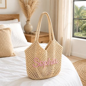 May include: A beige crochet tote bag with two long handles and an open-weave design. The front of the bag features the name Sophia embroidered in light pink yarn in a cursive script font, centered on the lower portion of the bag body.