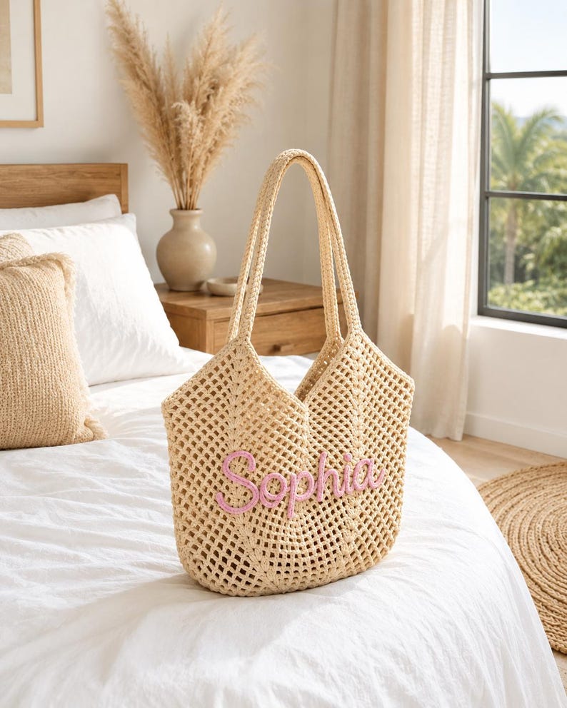 May include: A beige crochet tote bag with two long handles and an open-weave design. The front of the bag features the name Sophia embroidered in light pink yarn in a cursive script font, centered on the lower portion of the bag body.