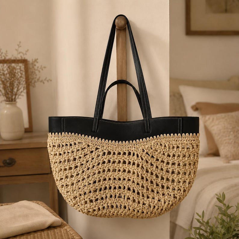 May include: A tote bag featuring a natural beige woven straw body with an open-knit pattern and a smooth black leather top panel. The bag includes two black leather shoulder straps with visible white stitching, creating a contrast between the textures.