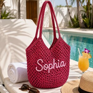 May include: A deep magenta crochet tote bag featuring the name Sophia embroidered in light pink script lettering on the front. The bag has an open-weave mesh construction with two long shoulder straps and a spacious, unlined interior compartment.