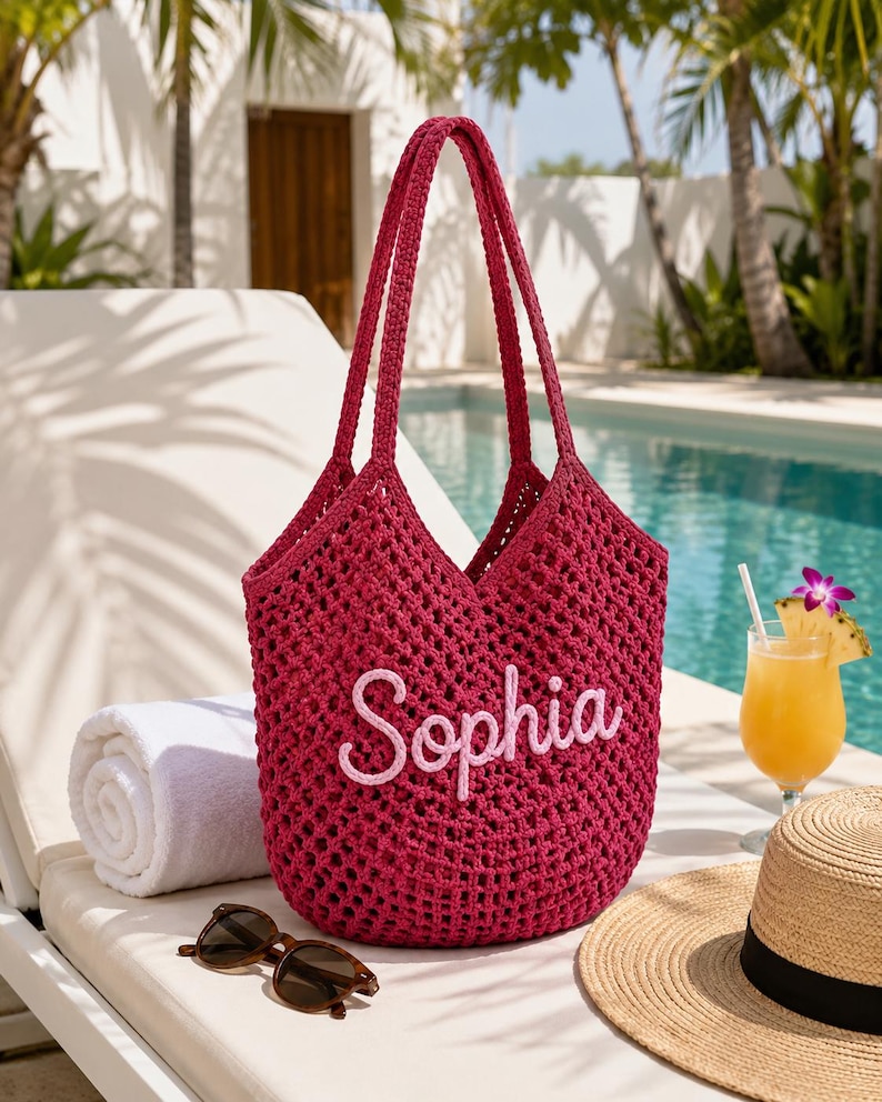 May include: A deep magenta crochet tote bag featuring the name Sophia embroidered in light pink script lettering on the front. The bag has an open-weave mesh construction with two long shoulder straps and a spacious, unlined interior compartment.