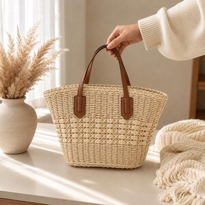 Pode incluir: Uma bolsa tote de palha tran&ccedil;ada em tom bege natural, com um padr&atilde;o de treli&ccedil;a texturizado na parte superior e duas al&ccedil;as de couro castanho fixadas com abas retangulares. A bolsa possui uma abertura larga e uma base estruturada.