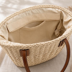 Pode incluir: Uma bolsa tote de palha tran&ccedil;ada com forro de tecido bege, apresentando um compartimento interno aberto com um bolso e duas al&ccedil;as de ombro de aspecto couro marrom fixadas com remendos retangulares na parte externa da bolsa.