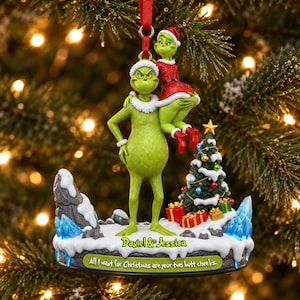 Peut inclure: Une décoration de Noël représentant un personnage vert tenant un plus petit personnage vert en robe rouge. La base affiche le texte 'Daniel & Jessica' et 'All I want for Christmas are your two butt cheeks.' Inclut un sapin décoré.