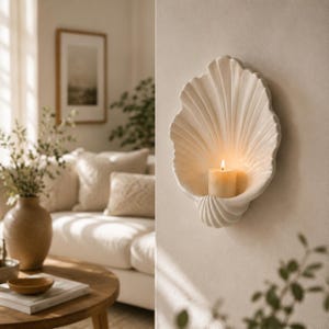 May include: A cream-colored ceramic wall sconce shaped like a large seashell, featuring a fluted design with a central indentation that holds a single lit pillar candle, creating a warm glow against the curved, textured surface of the shell.