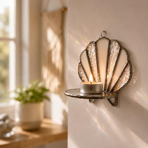 May include: A wall-mounted candle holder shaped like a seashell, crafted from iridescent stained glass with dark metal soldering. The piece features a small circular shelf holding a single lit tea light candle, creating a soft glow.