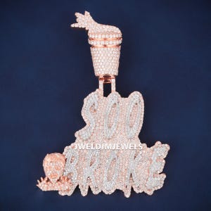 May include: A rose gold pendant featuring a cup with a hand gesture on top, connected to a large script text piece that reads "SOO BROKE" with the text "JWELDIMJEWELS" in the center. The entire piece is covered in small, clear, sparkling stones.