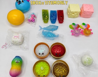 Pode incluir: Uma coleção de vários brinquedos squishy incluindo um cão azul, uma bola amarela, tubos com glitter, figuras de tubarão, blocos de queijo, peixes coloridos, recipientes redondos com rostos sorridentes e um amendoim arco-íris com o texto Surprise Box.