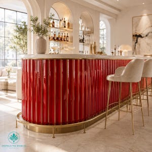 Op de afbeelding: Een gebogen bar met glanzende rode verticaal geribbelde tegels en gouden metalen accenten. De bar heeft een wit stenen blad en een gouden metalen voetsteun. Twee beige gestoffeerde barkrukken met gouden metalen poten staan ernaast.