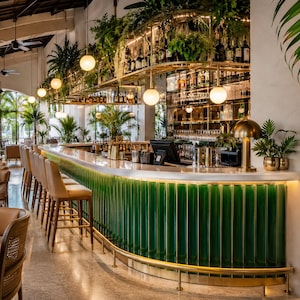 May include: A curved bar counter featuring vertical green glass panels and a gold metal trim. The bar is lined with light-coloured upholstered stools with wooden legs. Above the counter, a tiered gold shelf holds bottles and hanging green plants.