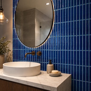 May include: A bathroom vanity area featuring a white circular vessel sink, a bronze faucet, and a soap dispenser with a bar of soap. The wall is covered in vertical, glossy, deep blue rectangular tiles, complemented by a round mirror with backlighting.