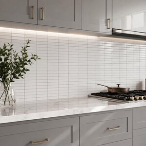 May include: A kitchen featuring gray shaker-style cabinets with metallic handles, a white marble countertop, and a white rectangular tile backsplash. A stainless steel pan sits on a black gas stovetop, and a glass vase holds green foliage.