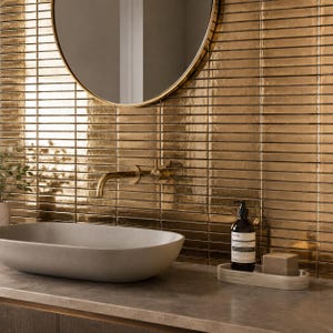May include: A bathroom vanity area featuring a wall covered in horizontal metallic gold rectangular tiles. A round mirror hangs above a wall-mounted brass faucet and a light gray oval basin. A dark soap bottle and a small block sit on a tray.