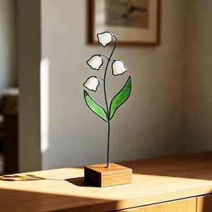Könnte beinhalten: Eine Skulptur aus Buntglas in Form eines Maiglöckchens mit drei weißen glockenförmigen Blüten und zwei grünen Blättern an einem dünnen schwarzen Metallstiel, montiert auf einem rechteckigen Holzsockel mit natürlicher Maserung.