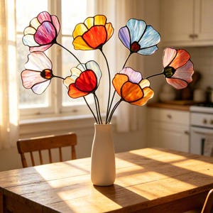 May include: A collection of seven stained glass flowers with thin black metal stems arranged in a tall, smooth, matte white ceramic vase. Each flower features translucent, multi-coloured glass petals in shades of pink, yellow, orange, and blue.