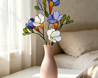 May include: A light pink ceramic vase containing a bouquet of stained glass flowers. The flowers feature petals in shades of white, blue, and purple with orange centers, connected by thin dark metal lines, accompanied by small green glass leaves.
