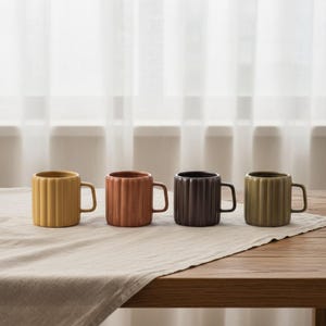 May include: Four ceramic mugs arranged in a row, each featuring a vertical ribbed texture and a side handle. The mugs are colored in mustard yellow, terracotta orange, dark brown, and olive green, all with a consistent cylindrical shape.