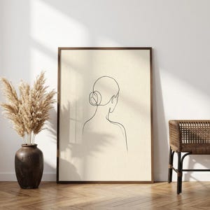 May include: A framed wall art print featuring a black single-line drawing of a person seen from behind with hair in a bun. The artwork is set against a light beige background with a subtle speckled texture, enclosed in a dark brown wooden frame.
