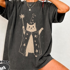 May include: A charcoal gray short-sleeve t-shirt featuring a graphic print of a cat dressed as a wizard, wearing a star-patterned robe and hat while holding a star-topped wand. The shirt has a loose, relaxed fit with a crew neckline.
