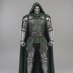 May include: A collectible action figure of a character wearing a metallic silver mask and full suit of armor, featuring a dark green hooded cloak, a textured tunic, a black belt with a circular buckle, and silver gauntlets and greaves.