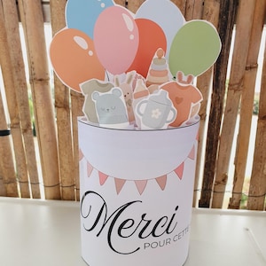 Peut inclure: Une carte cylindrique avec le texte "Merci POUR CETTE" et une guirlande rose. Le sommet présente des découpes de ballons colorés, un body, une tête d'ours, un lapin, un biberon, un gobelet à bec et un jouet empilable aux tons pastel.