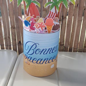 Peut inclure: Une décoration cylindrique en papier illustrant une scène de plage avec le texte Bonne vacances. La base représente du sable et des coquillages, tandis que le sommet présente des découpes de palmiers, une planche de surf, un flamant rose.