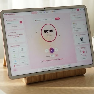 Può includere: Un tablet posizionato su un supporto in legno, che mostra un'applicazione timer per lo studio con un conto alla rovescia di 90:00, un pulsante play e varie icone di navigazione su un'interfaccia di colore rosa chiaro e bianco.