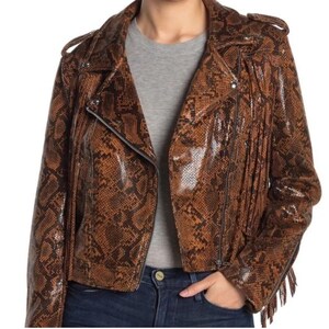 Puede incluir: Una chaqueta motera con estampado de piel de serpiente en tonos marrón y negro, con solapa de muesca, mangas largas con flecos, charreteras en los hombros y cierre de cremallera frontal. La prenda tiene un acabado brillante y un t-shirt gris.