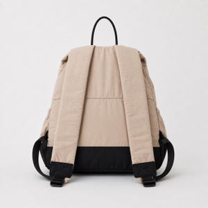 May include: A beige backpack with a black base and black shoulder straps. The backpack features a top handle, padded shoulder straps with adjustable black buckles, and a horizontal seam across the back panel, showcasing a two-tone colour design.