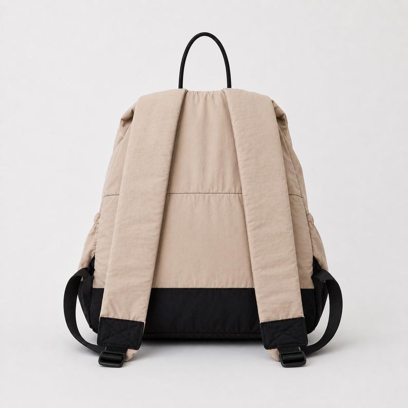 May include: A beige backpack with a black base and black shoulder straps. The backpack features a top handle, padded shoulder straps with adjustable black buckles, and a horizontal seam across the back panel, showcasing a two-tone colour design.