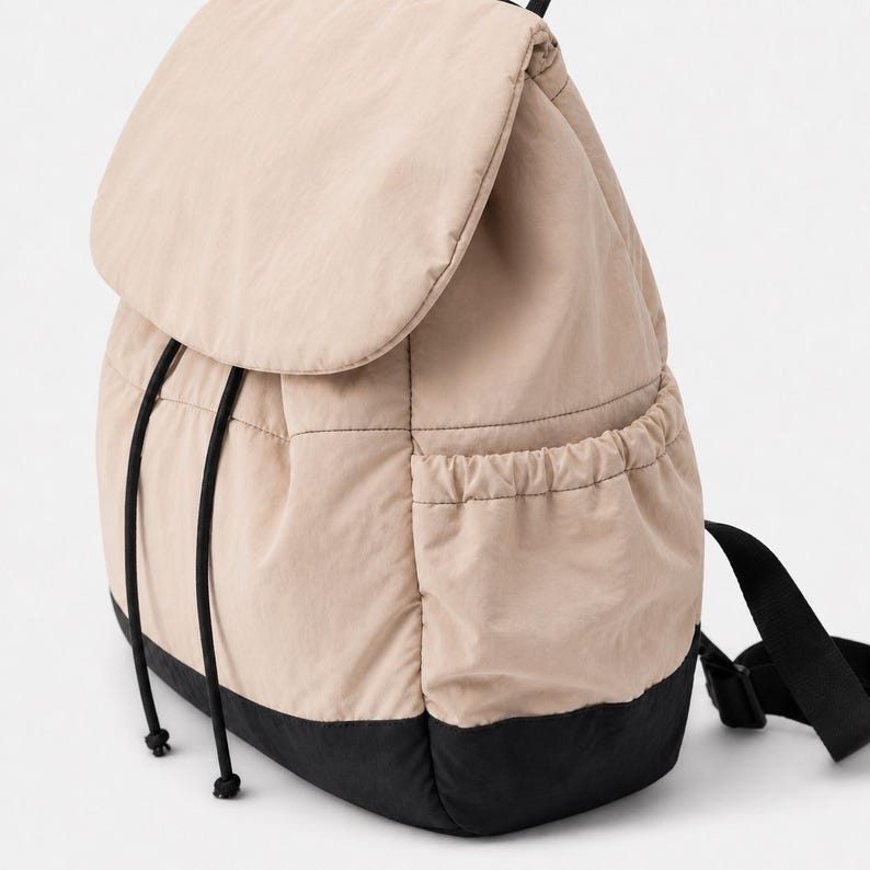 May include: A beige quilted backpack featuring a large top flap, black drawstring cords, and a black base. The bag includes side pockets with elasticated openings and black adjustable shoulder straps attached to the back panel.