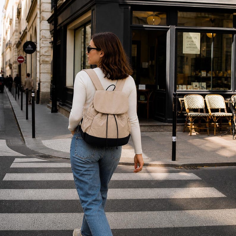 May include: A beige backpack with a black base, featuring a top flap, a drawstring closure, and two shoulder straps. The bag has side pockets and is constructed from a fabric material, showcasing a two-tone design with black cord accents.