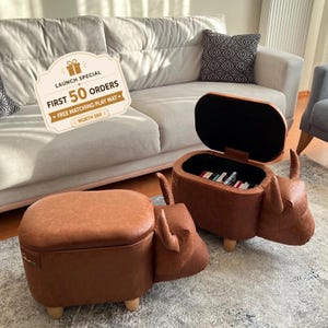 Op de afbeelding: Twee bruine, diervormige opberghockers met gladde kunstlederen oppervlakken en kleine houten pootjes. De ene hocker is gesloten, terwijl de andere open is en een zwart interieur vol boeken onthult. Een bordje vermeldt: LAUNCH SPECIAL FIRST 50 ORDERS FREE MATCHING PLAY MAT WORTH $80.