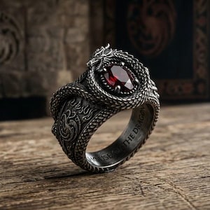 May include: A silver-toned metal ring featuring a coiled dragon design with textured scales and a central oval-cut deep red gemstone. The band is engraved with the text HOUSE OF THE DRAGON and features intricate flame patterns along the sides.
