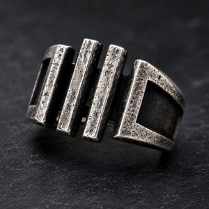 Puede incluir: Un anillo de metal en tono plateado con un acabado texturizado y desgastado. El diseño consta de cuatro barras verticales de diferentes anchos conectadas por una banda horizontal, creando una estructura geométrica abierta con áreas oscuras.