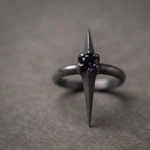 May include: A dark gray metal ring featuring a pointed, star-like setting with a single, round, deep purple gemstone held in place by six prongs. The band has a matte, textured finish and the central design extends vertically above the ring band.