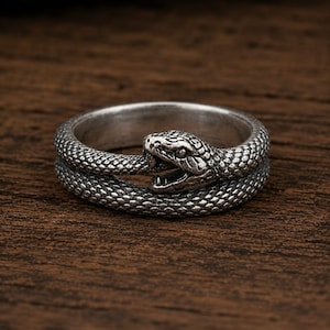 May include: A silver-coloured metal ring featuring a coiled snake design. The snake's body forms the band of the ring, with the textured scales wrapping around to meet the head, which has an open mouth showing small teeth and detailed eye markings.