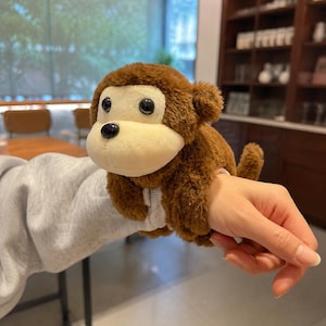 May include: A plush monkey toy designed as a wrist accessory, featuring brown fur, a cream-colored face, and black plastic eyes. The toy has long arms that wrap around the wrist, with a small tail visible on the side, creating a soft, wearable item.