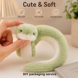 Puede incluir: Un juguete de peluche con forma de criatura curva con pelaje verde claro en la parte posterior y blanco en la parte frontal. El juguete presenta pequeños ojos oscuros y una textura suave. El texto en la imagen incluye "Cute & Soft", "Ultra Soft", "Skin Friendly", "Adorable Design" y "DIY packaging service".