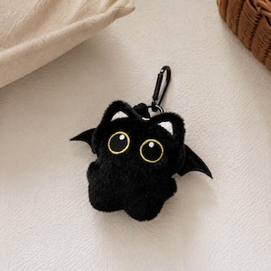 May include: A plush black keychain shaped like a bat with large yellow-rimmed eyes, silver-colored inner ears, and small black wings. The soft, fuzzy character features a black metal lobster clasp attached to the top for easy hanging and secure use.