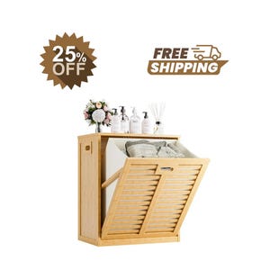 May include: A light brown bamboo laundry hamper with a tilt-out front panel featuring horizontal slats and a white interior liner. The top surface holds a small bouquet of flowers, four clear pump bottles, and a glass jar with reed sticks.