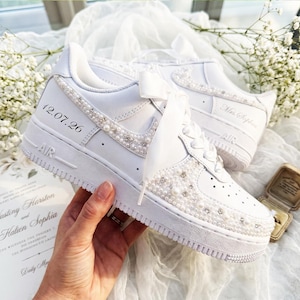 Puede incluir: Una zapatilla blanca con cordones de cinta de satén, adornos de perlas y pedrería en el lateral y la puntera, y el texto 12.07.26 impreso en el talón. El calzado incluye una suela de goma gruesa y otra zapatilla blanca a juego detrás.