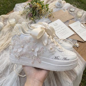 Può includere: Una sneaker bianca con lacci in nastro di raso, applicazioni in pizzo floreale e decorazioni di perle. Il lato della scarpa mostra il testo Mis. Alice in un carattere corsivo nero. La zona del tallone è decorata con piccole perle bianche.
