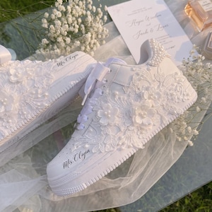 May include: A pair of white sneakers featuring intricate floral lace appliques, pearl embellishments, and wide satin ribbon laces. The side of the shoe is customized with the black cursive text "Mrs. Olyia" printed on the leather surface.