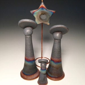 Può includere: Un set di tre portacandele in ceramica di varie dimensioni, con un ornamento a forma di stella sospeso sopra. I portacandele sono grigi con bande iridescenti di colore e la stella ha una finitura simile. Il design include un elemento in filo di rame.
