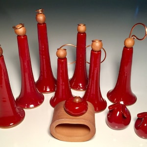 以下が含まれることがあります： 深紅色の釉薬を施した手作りの陶器のコレクション。木製の球体と銅線アクセントをあしらった背の高いトランペット型のフォルム、小さな丸いオブジェ、テラコッタ製のナプキンリングが含まれています。白い表面に配置されています。
