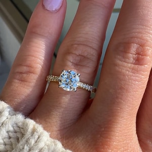 May include: A gold ring featuring a large, round, brilliant-cut center stone held by four prongs, with a band encrusted with small, sparkling pavé-set diamonds extending along the sides of the shank, creating a bright and reflective appearance.