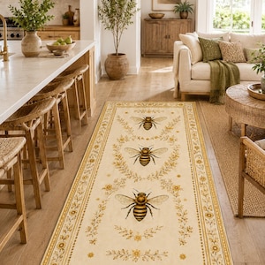 Op de afbeelding: Een rechthoekig vloerkleed met een crèmekleurige achtergrond en een herhalend patroon van drie centrale honingbijen, omringd door delicate gouden bloemenkransen en een decoratieve rand van kleine gele bloemen en rankmotieven.