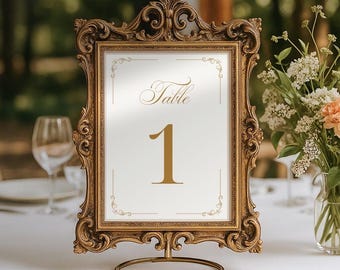 May include: A vertical rectangular table number sign featuring the word Table and the number 1 in gold script and serif font. The sign is encased in an ornate, antique gold-colored frame with intricate scrollwork and rests on a circular metal stand.